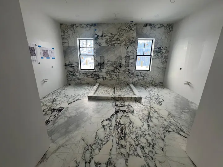 Primary bathroom with bookmatched marble-style walls and floor, before fixtures